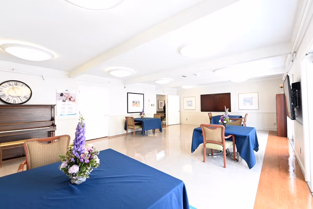 A bright and clean dining room with several tables covered in blue tablecloths, each adorned with a floral centerpiece. The room features a piano against one wall, a large clock, framed artwork, and a television mounted on the opposite wall. Chairs are neatly arranged around the tables, and the floor is a combination of light-colored tiles and wood.