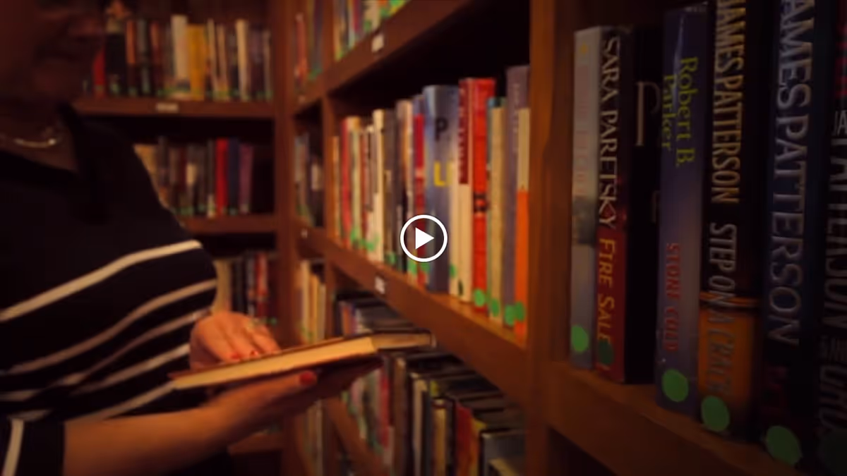 A person wearing a striped shirt is selecting a book from a wooden bookshelf filled with various books in a library or reading area.