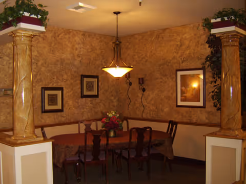 Cozy dining room with a round table covered by a tablecloth, several chairs, decorative columns, a hanging light fixture, framed artwork and plants.