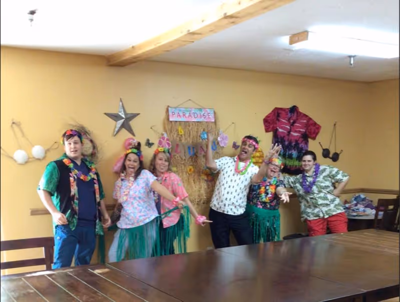 Six people dressed in colorful Hawaiian-themed outfits and leis posing cheerfully in a room with yellow walls. Behind them is a decorative wall hanging with the word 'PARADISE' and tropical decorations. There is a wooden table in the foreground and some chairs along the walls.