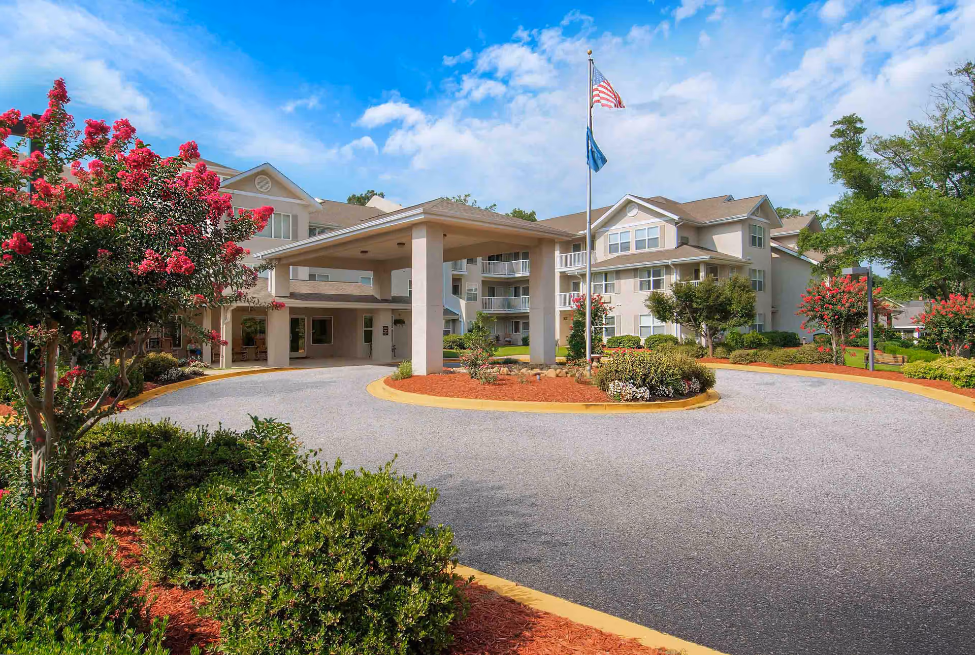 Exterior view of TerraBella Greenville senior living facility showing a circular driveway with a covered entrance, landscaped bushes and flowering trees, and an American flag flying on a flagpole under a partly cloudy blue sky.