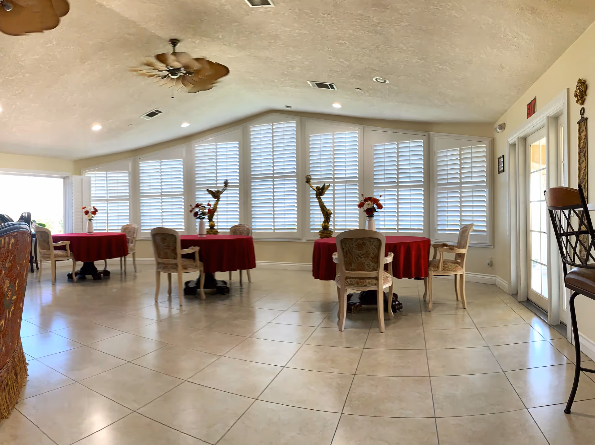 A bright dining area with several round tables covered in red tablecloths, each surrounded by ornate chairs. The room features large windows with white plantation shutters, two decorative golden eagle statues on the windowsill, and ceiling fans with leaf-shaped blades. The floor is tiled, and there is a door with glass panels on the right side.