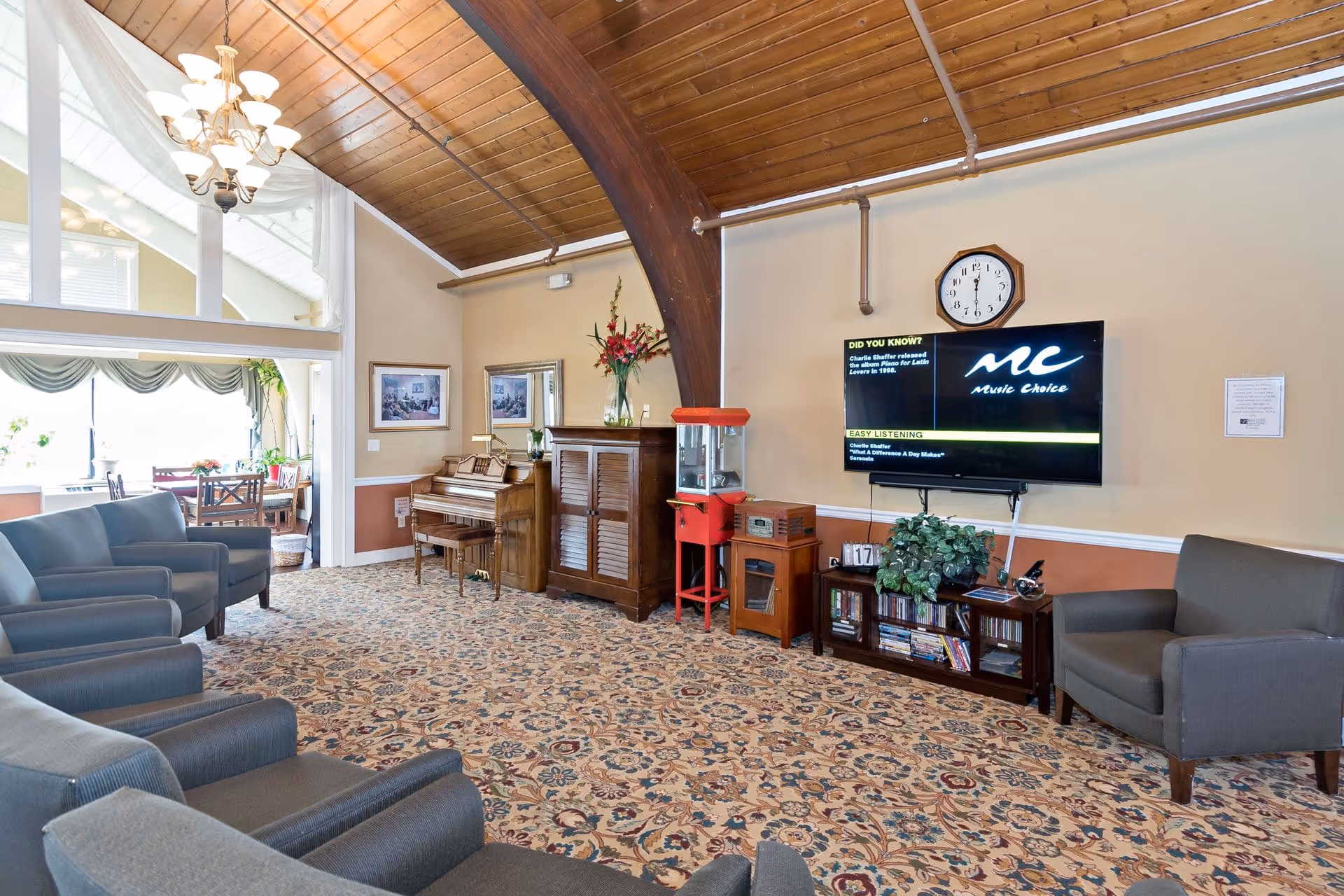 Bright communal living area with upholstered chairs, a piano, wall-mounted TV, popcorn machine and a dining nook under a wood-paneled vaulted ceiling.