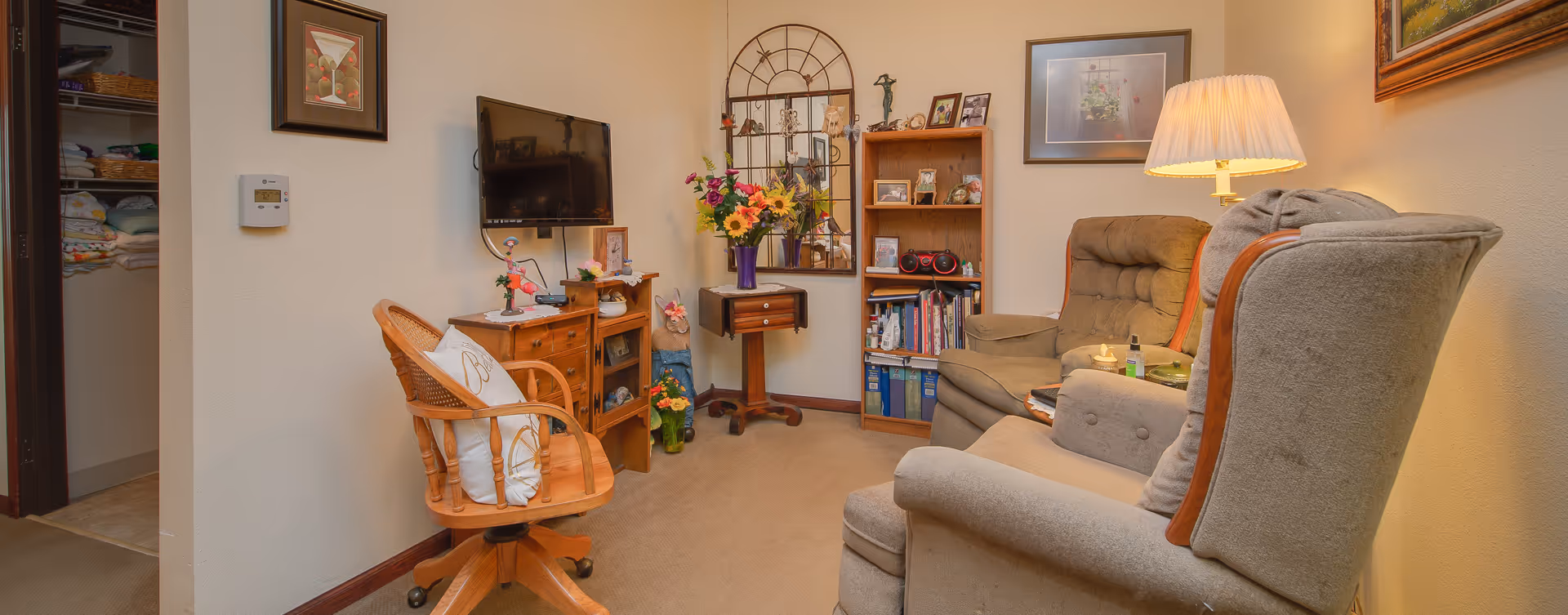 A cozy living room with two cushioned armchairs and a wooden swivel chair with a white pillow. There is a wooden cabinet with a flat-screen TV mounted above it, a small wooden table with a vase of colorful flowers, and a wooden bookshelf filled with books, framed photos, and a radio. A lamp with a pleated shade is turned on, casting warm light. The walls are decorated with framed pictures and a decorative mirror. An open doorway reveals shelves stocked with towels and linens.