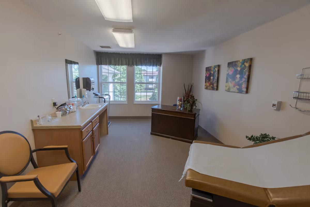 A medical examination room with a padded examination table covered with white paper on the right, a wooden desk with a floral arrangement and decorative balls in a glass vase near a window with green curtains, a beige chair with wooden arms on the left, a countertop with a sink, soap dispenser, and medical supplies, and two colorful abstract paintings on the wall.