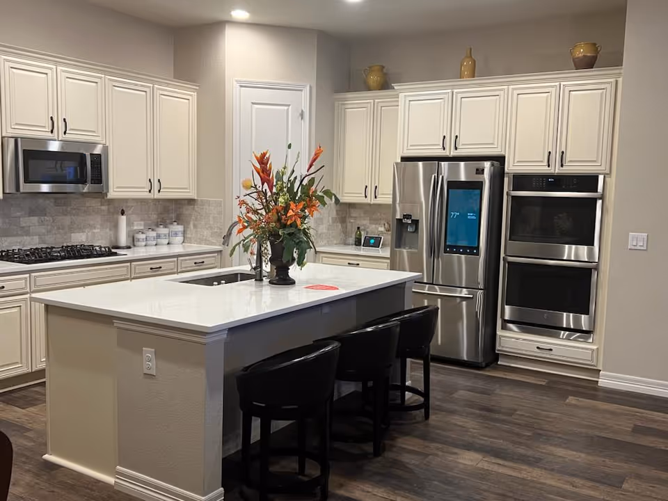 Modern kitchen with white cabinetry, a large island with a sink and three black chairs, stainless steel appliances including a refrigerator with a digital screen, double oven, and microwave. There is a vase with orange and green flowers on the island and decorative pottery on top of the cabinets.
