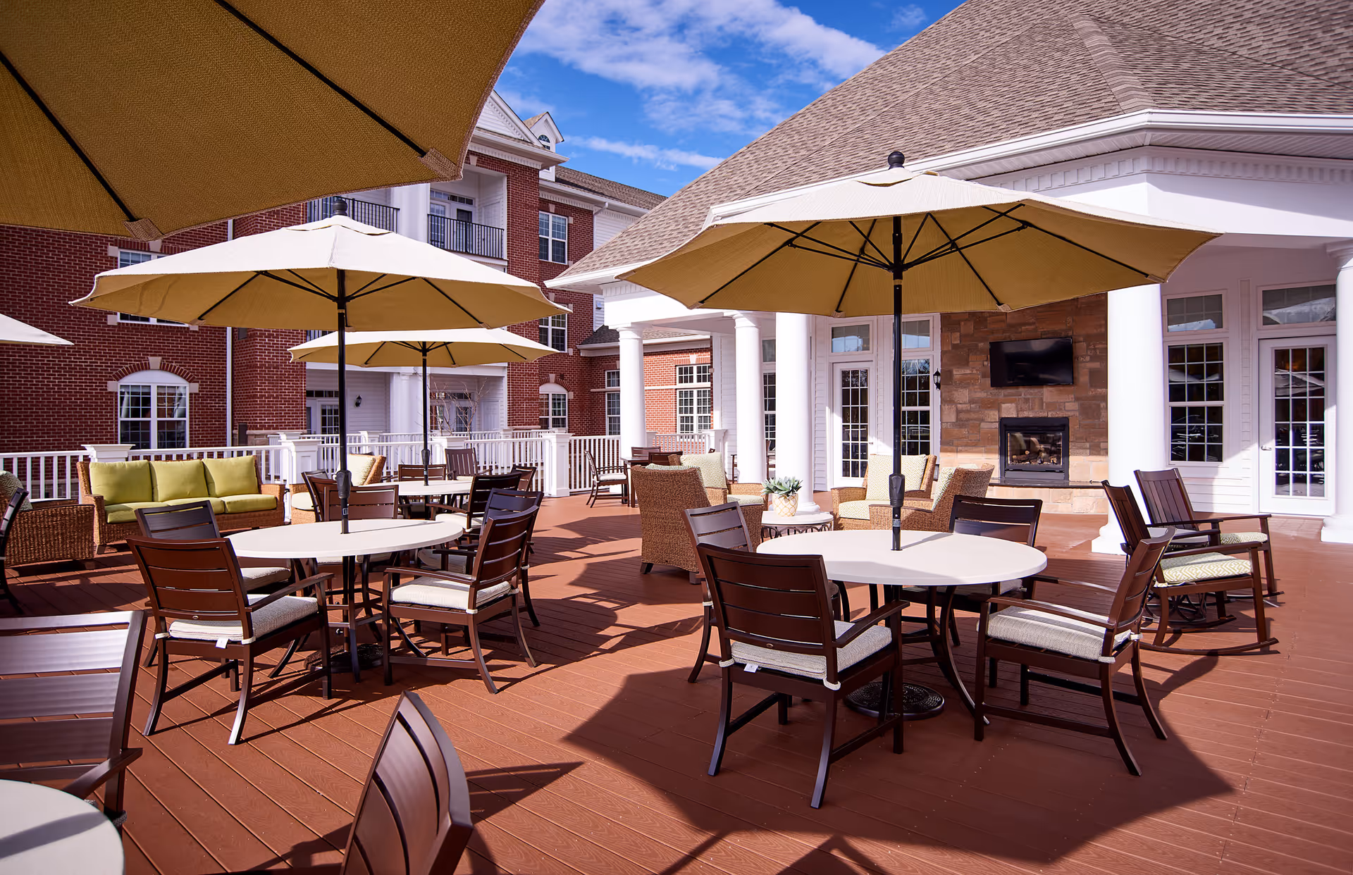 Outdoor patio area with multiple round tables and chairs, each table shaded by large beige umbrellas. The patio has a reddish-brown wooden floor and is adjacent to a building with white columns, large windows, and a mounted TV above a stone fireplace. The sky is clear with a few clouds.