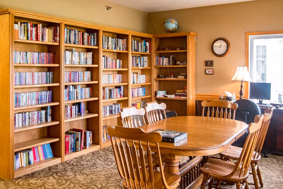 A cozy library room with wooden bookshelves filled with books along the wall, a wooden table surrounded by six wooden chairs in the center, a globe on top of a bookshelf, a wall clock, a desk with a computer monitor, and a table lamp near a window.