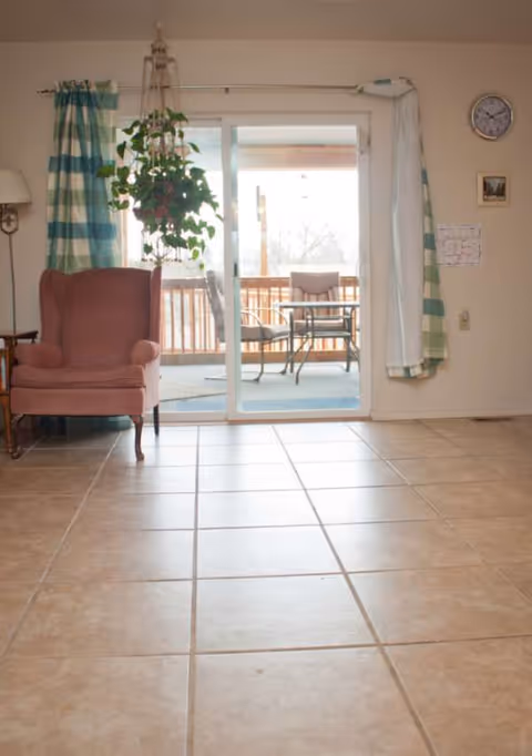 Empty living room with tile floor, pink chair, hanging plant, and sliding glass door.