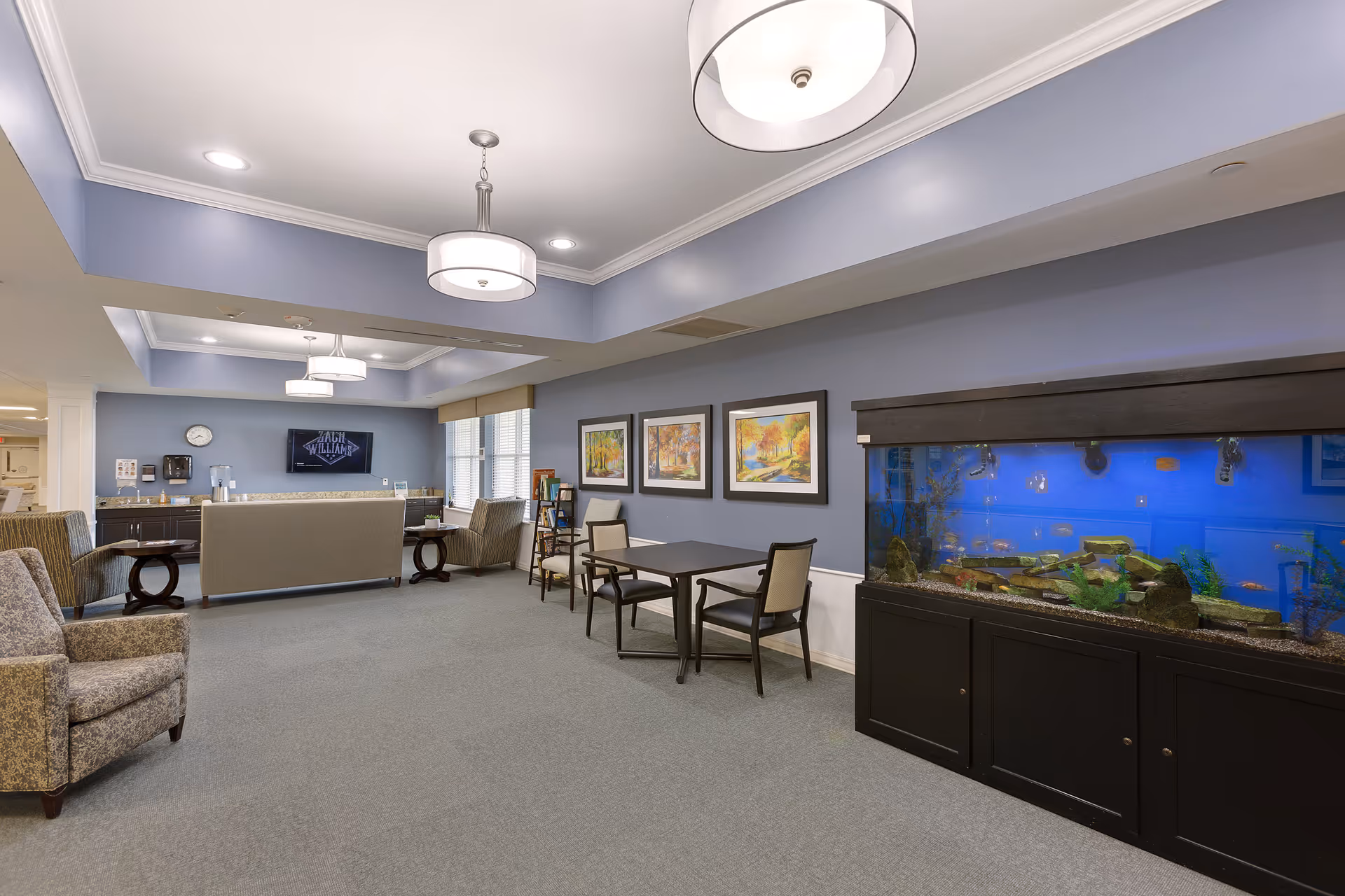 A spacious, well-lit common area with light blue walls and gray carpet. The room features comfortable armchairs, a sofa facing a wall-mounted TV, a large aquarium with colorful fish, and a table with chairs. Three framed landscape paintings hang on the wall above the table. Ceiling lights provide ample illumination.