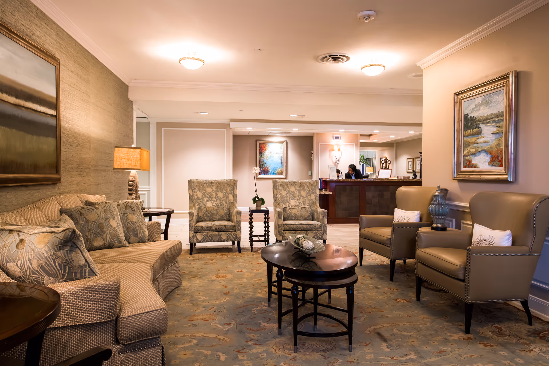 A cozy and well-lit living room area in a senior living facility with a beige sofa, patterned armchairs, two leather armchairs with white pillows, a round coffee table, and framed paintings on the walls. In the background, a reception desk with a staff member is visible.