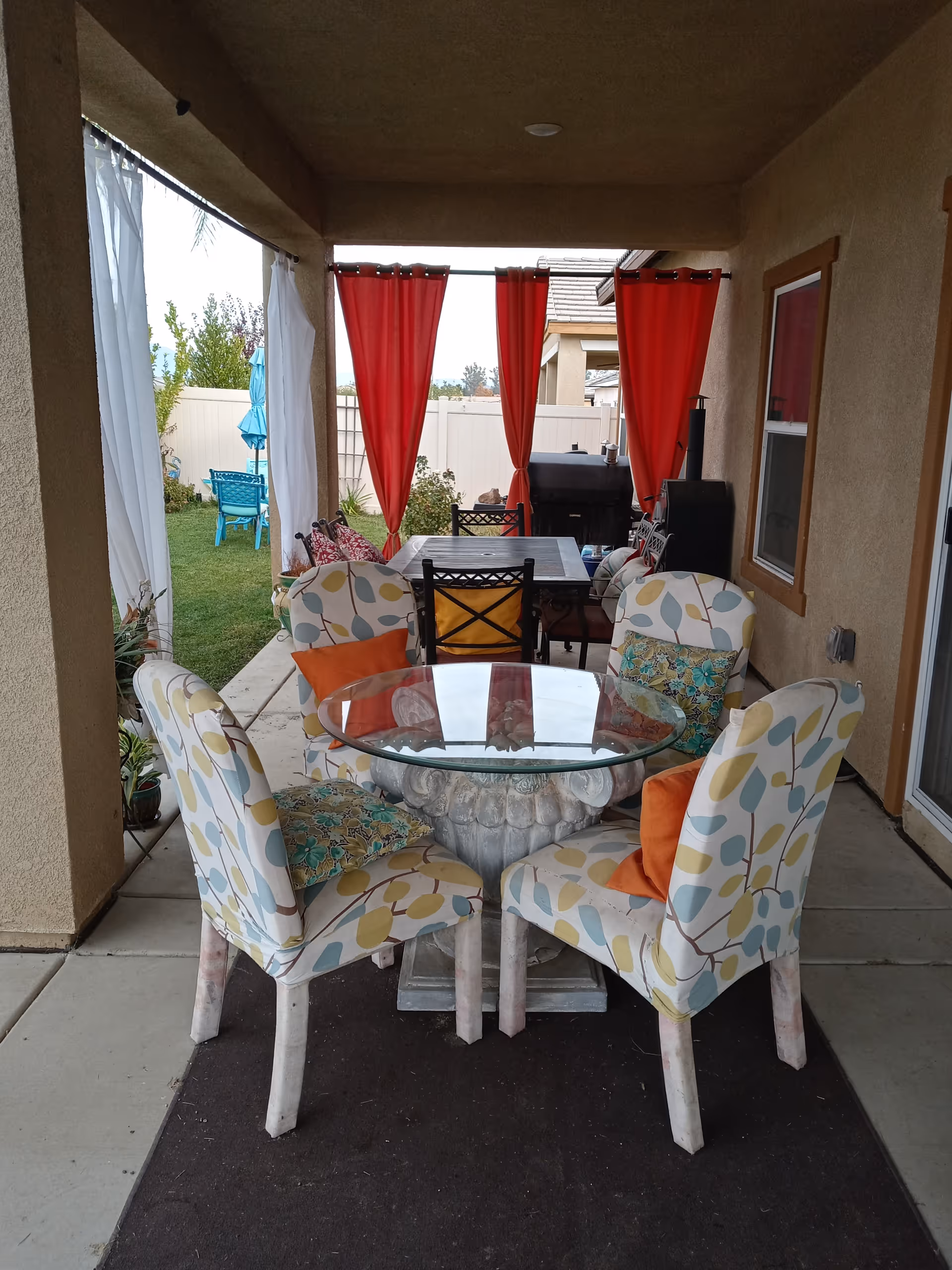 Covered outdoor patio area with a round glass-top table surrounded by four patterned chairs with orange and floral cushions. Behind the table is a rectangular dining table with chairs, red curtains hanging from the ceiling, and a barbecue grill. The patio opens to a grassy yard with blue outdoor furniture and an umbrella.