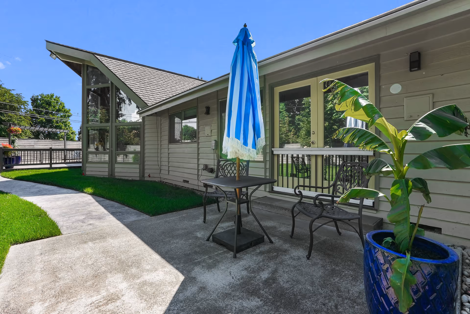 Outdoor patio area with a small black metal table and two matching chairs. A blue and white striped umbrella is closed and positioned in the center of the table. There is a large potted plant with broad green leaves in a blue ceramic pot on the right side. The patio is adjacent to a light gray building with large windows and a door. A curved concrete pathway runs alongside a well-maintained green lawn under a clear blue sky.
