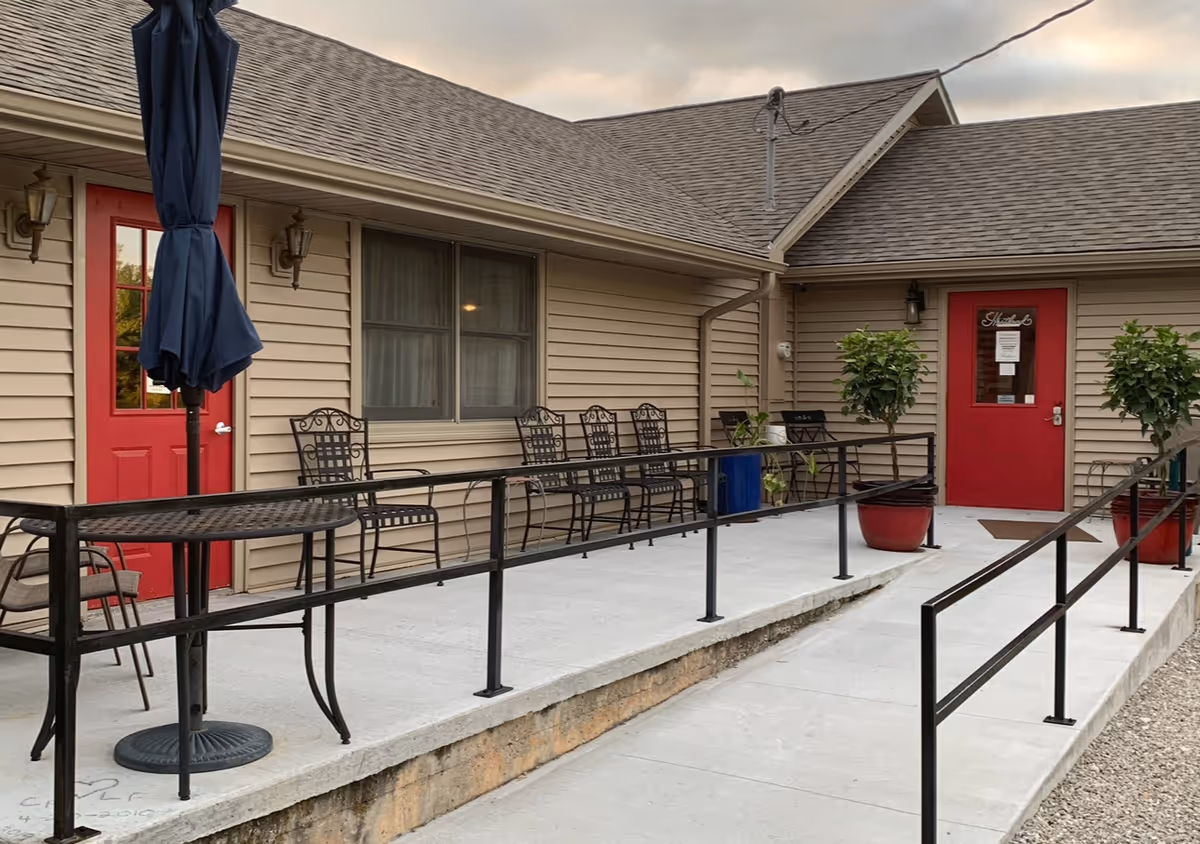 Outdoor patio area at Hartland Residential Care Center with beige siding, two red doors, black metal chairs lined up against the wall, a black metal table with a closed navy blue umbrella, and two large potted plants near the entrance. The patio has a concrete ramp with black handrails.