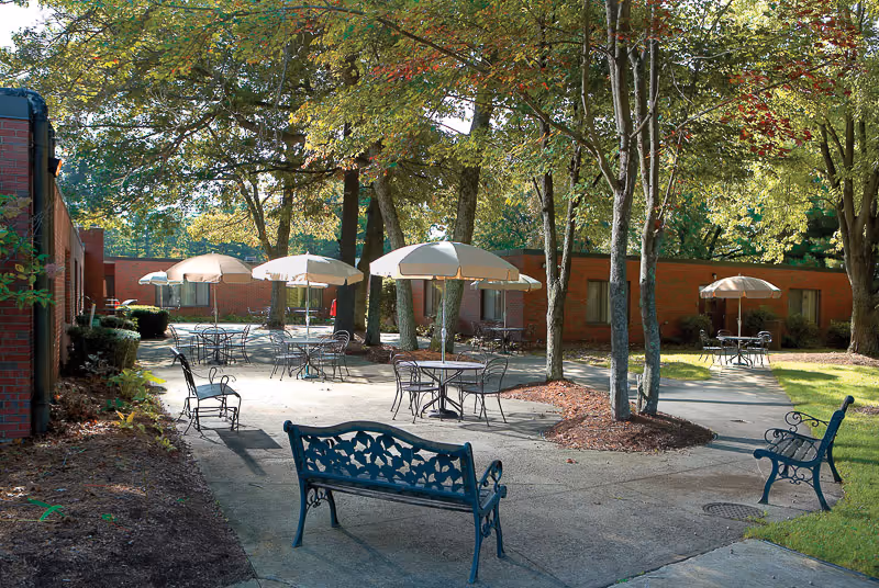 Outdoor patio area with metal tables and chairs under large umbrellas surrounded by trees and grass, adjacent to a single-story brick building.