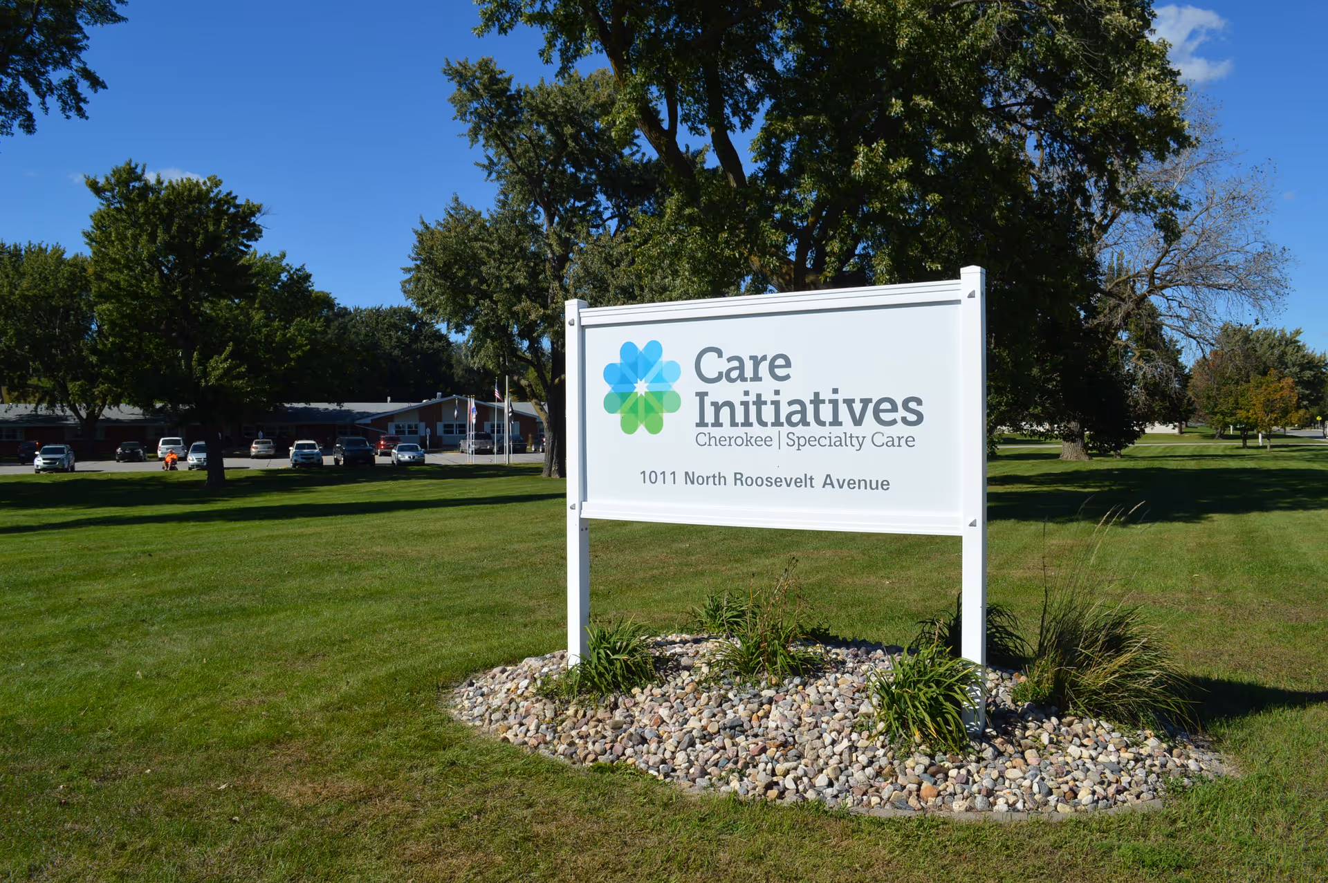 A white sign with the logo and text 'Care Initiatives Cherokee Specialty Care 1011 North Roosevelt Avenue' is displayed on a grassy area with trees and a building with parked cars in the background under a clear blue sky.