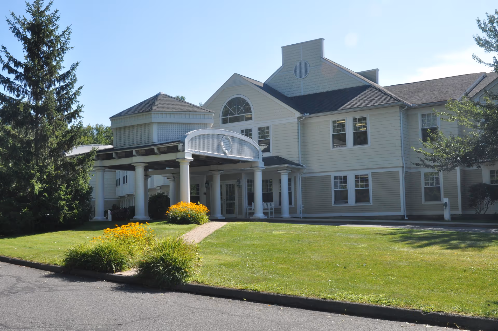 Exterior view of The Arbors at Amherst Assisted Living and Memory Care building with a covered entrance supported by white columns, surrounded by green lawn, trees, and yellow flowers under a clear blue sky.