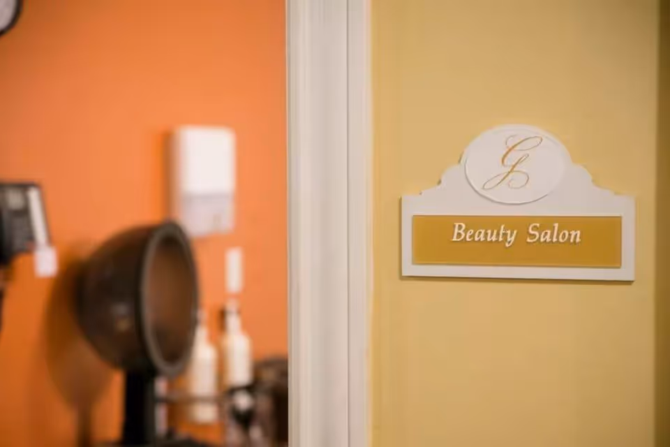 A close-up view of a wall sign that reads 'Beauty Salon' with a stylized 'G' above it, mounted on a yellow wall. In the background, there is an orange wall with a hair dryer and some bottles, indicating a salon setting.