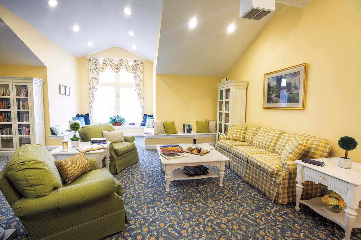 A cozy living room with yellow walls and a patterned blue carpet. The room features a green armchair, a yellow plaid sofa, a white coffee table with books and a tray of snacks, and white side tables with small potted plants. There is a large window with floral curtains and a built-in bench with cushions beneath it. A white cabinet filled with books is visible on the left side, and a framed picture hangs on the wall above the sofa.