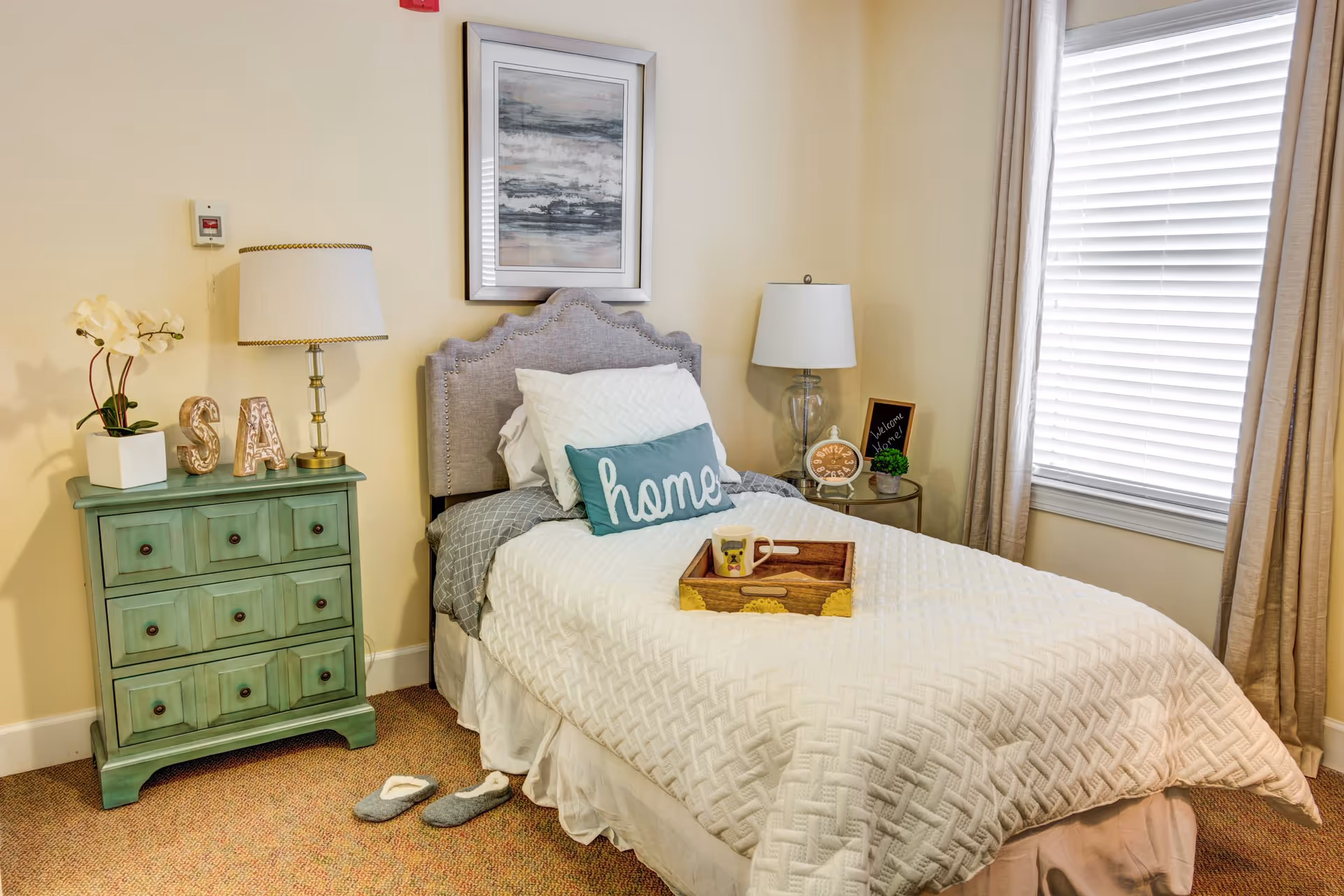 Cozy single bedroom with a neatly made bed, a 'home' pillow, bedside tables and lamps, a green dresser, and a window with blinds.