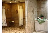 Tiled communal bathroom with two curtained shower stalls, a shower chair, and a sink vanity with towels and a plant.