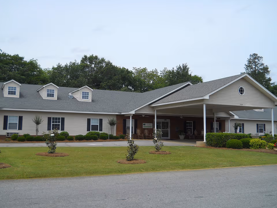 Exterior view of Century Pines Senior Retire facility showing a single-story building with a covered entrance, multiple windows with black shutters, small trees and shrubs in front, and a paved driveway.