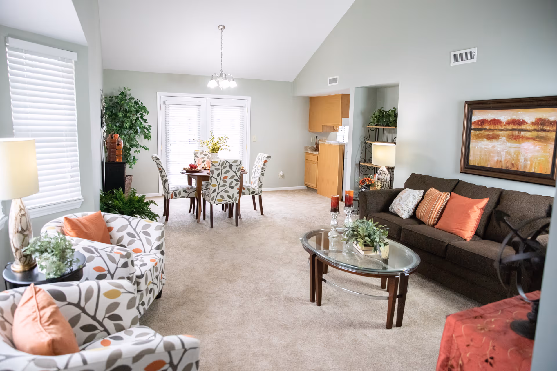 A bright and spacious living room and dining area with light gray walls and beige carpet. The living room features a dark brown sofa with orange and patterned throw pillows, two patterned armchairs with orange pillows, a glass coffee table with decorative plants and candles, and a side table with a lamp. The dining area has a round wooden table with four patterned chairs and a vase with flowers. There are large windows with white blinds and a door leading outside. The kitchen is partially visible with wooden cabinets.