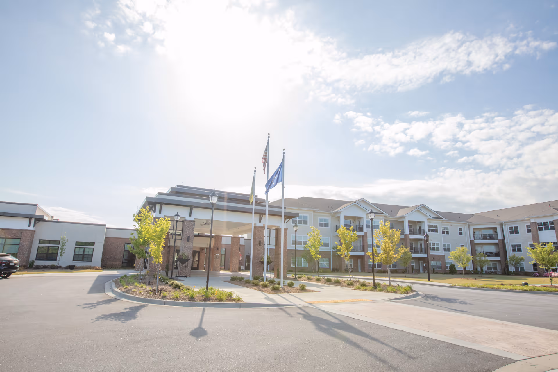 Exterior view of a large, modern senior living facility building with three stories, multiple windows, and balconies. The entrance features a covered drop-off area with three flagpoles flying flags. The surrounding area includes a paved driveway, landscaped greenery, and young trees under a partly cloudy sky with the sun shining brightly.