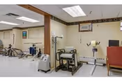 Interior view of a rehabilitation center room with exercise equipment including stationary bikes and other physical therapy machines. The room has white walls, a tiled floor, fluorescent ceiling lights, and a window air conditioning unit. There is a red chair visible on the right side.
