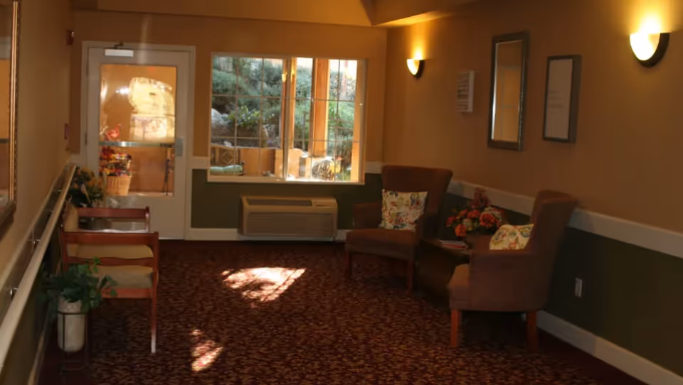 A cozy interior hallway area with patterned carpet, two upholstered chairs with floral cushions, a small table with a flower arrangement, a window showing an outdoor patio, and a door with a glass panel. The walls are painted beige and green with wall-mounted lights providing warm illumination.