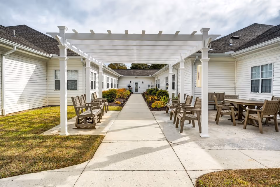 Outdoor courtyard area at Bridgewood Gardens featuring a white pergola over a concrete walkway, wooden chairs and tables arranged on either side, surrounded by grass and landscaping with bushes and plants, and white building walls with windows in the background under a partly cloudy sky.