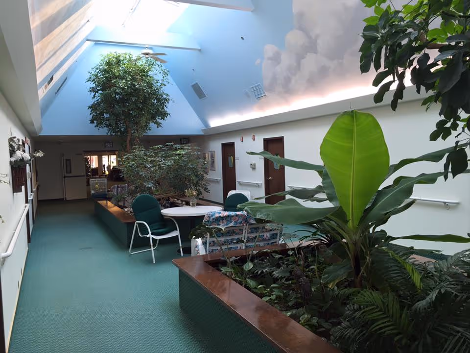 Indoor hallway area with green carpet and large planter boxes filled with various green plants and small trees. There are chairs and a round table near the plants. The ceiling is high and slanted with skylights allowing natural light to brighten the space. Doors line the walls, and handrails run along both sides of the hallway.