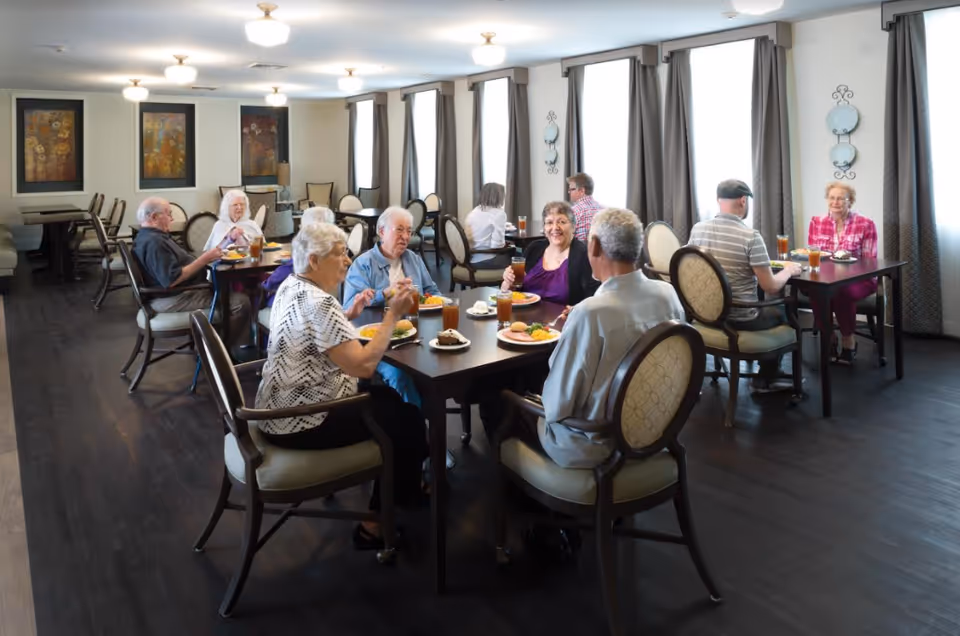 Elderly residents seated at tables eating and chatting in a bright communal dining room.