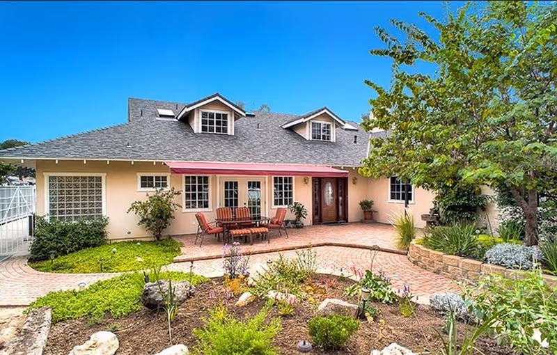 Exterior view of a single-story house with a gray shingled roof and beige walls. The front patio has a red awning, outdoor seating with chairs and a table, and is surrounded by a landscaped garden with various plants, shrubs, and a tree on the right side. The sky is clear and blue.