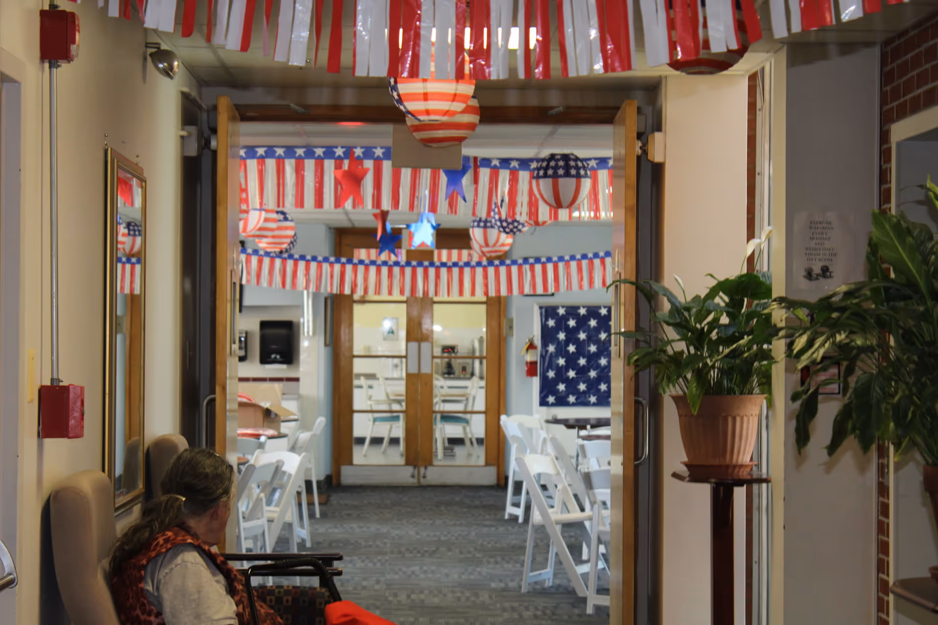 Interior view of a senior living facility hallway decorated with red, white, and blue patriotic banners and hanging lanterns. White folding chairs and tables are arranged in the adjoining room. A person in a wheelchair is seated in the foreground on the left side, and potted plants are visible on the right.