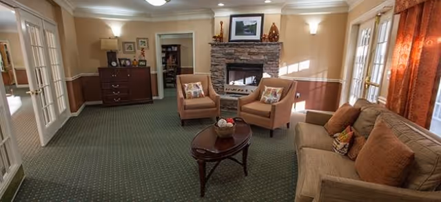 A cozy living room with a green carpet, a stone fireplace with a framed picture above it, two beige armchairs with decorative pillows, a brown sofa with cushions, a wooden coffee table with a decorative bowl, a wooden dresser with a lamp and framed photos, and large windows with orange curtains letting in natural light.