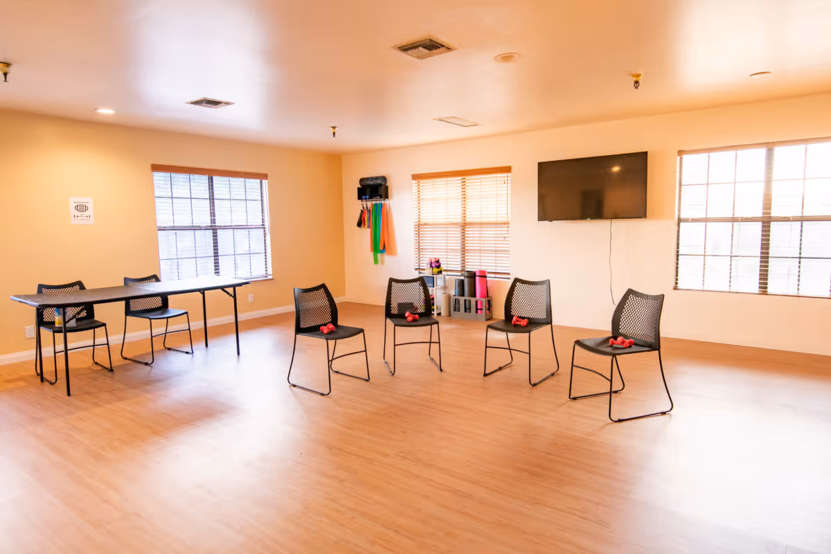 A bright room with wooden flooring and beige walls featuring four black chairs arranged in a semi-circle, each with a pair of small red dumbbells on the seat. There is a long black table with two chairs on the left side, three large windows with blinds, a wall-mounted flat screen TV, and various exercise equipment such as resistance bands and yoga mats stored on shelves and hooks.