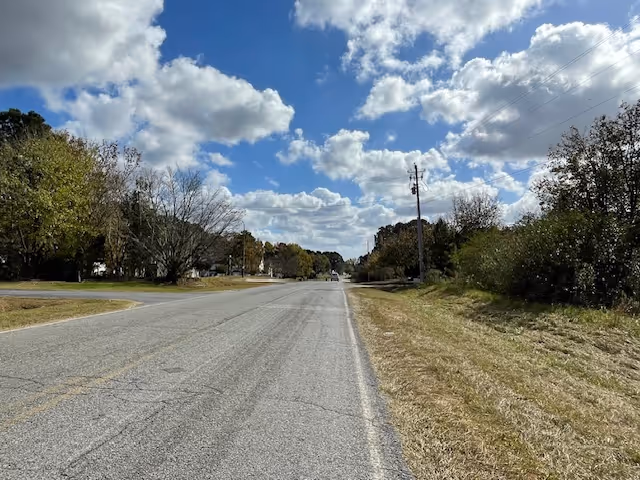 A two-lane rural road with grassy shoulders and trees under a partly cloudy blue sky.