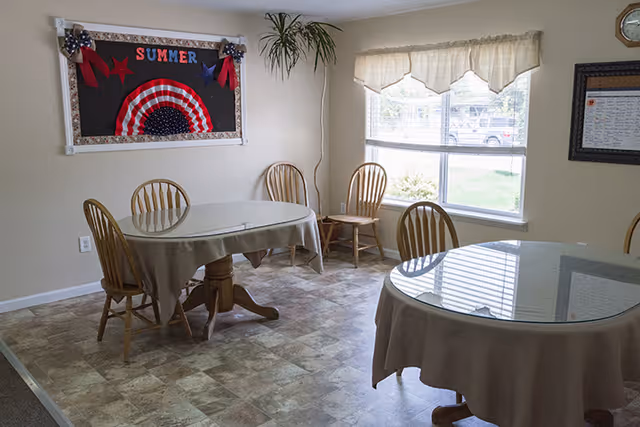 Bright dining room with round tables covered in tablecloths, wooden chairs, a window with a valance, and a summer-themed bulletin board on the wall.