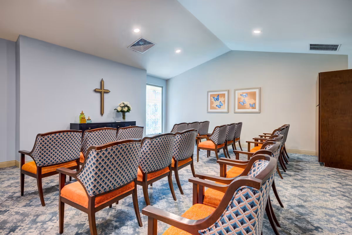 A small chapel or meditation room with rows of wooden chairs featuring orange cushions and patterned backs, a cross mounted on the wall above a cabinet with flowers and decorative items, two framed butterfly pictures on the opposite wall, and a window letting in natural light.