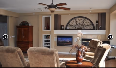 A cozy living room with beige walls and carpet, featuring a ceiling fan, a wooden cabinet, built-in shelves with decorative items, a wall-mounted TV above a fireplace, and several comfortable brown upholstered chairs arranged around a small round wooden table with a vase of flowers.