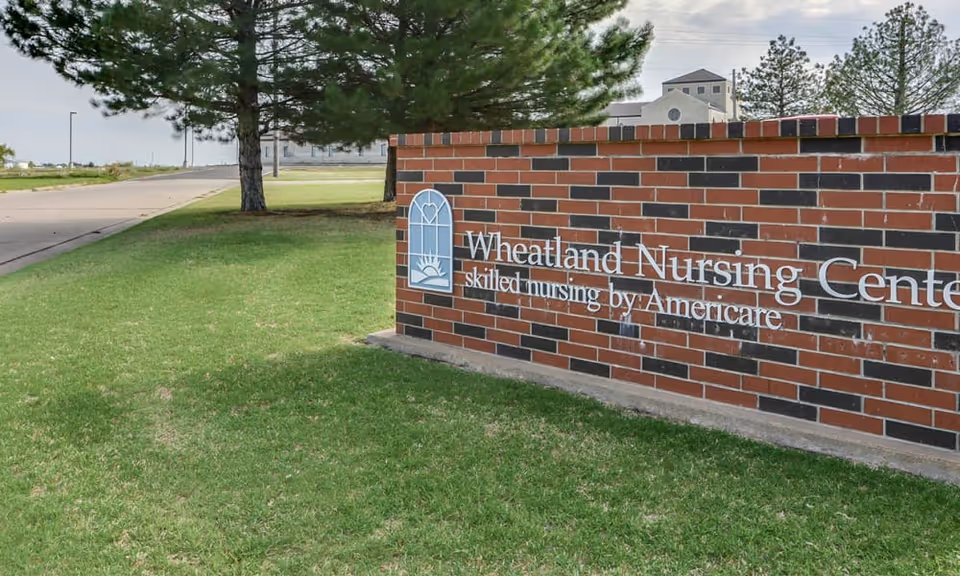 A brick sign for Wheatland Nursing Center with the text 'skilled nursing by Americare' on a grassy area next to a paved road, with trees and a building visible in the background under a partly cloudy sky.
