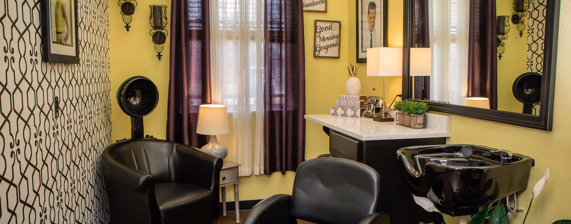 Small salon styling area with black chairs, a hood hair dryer, a wash basin, mirror, lamp, and patterned wallpaper.