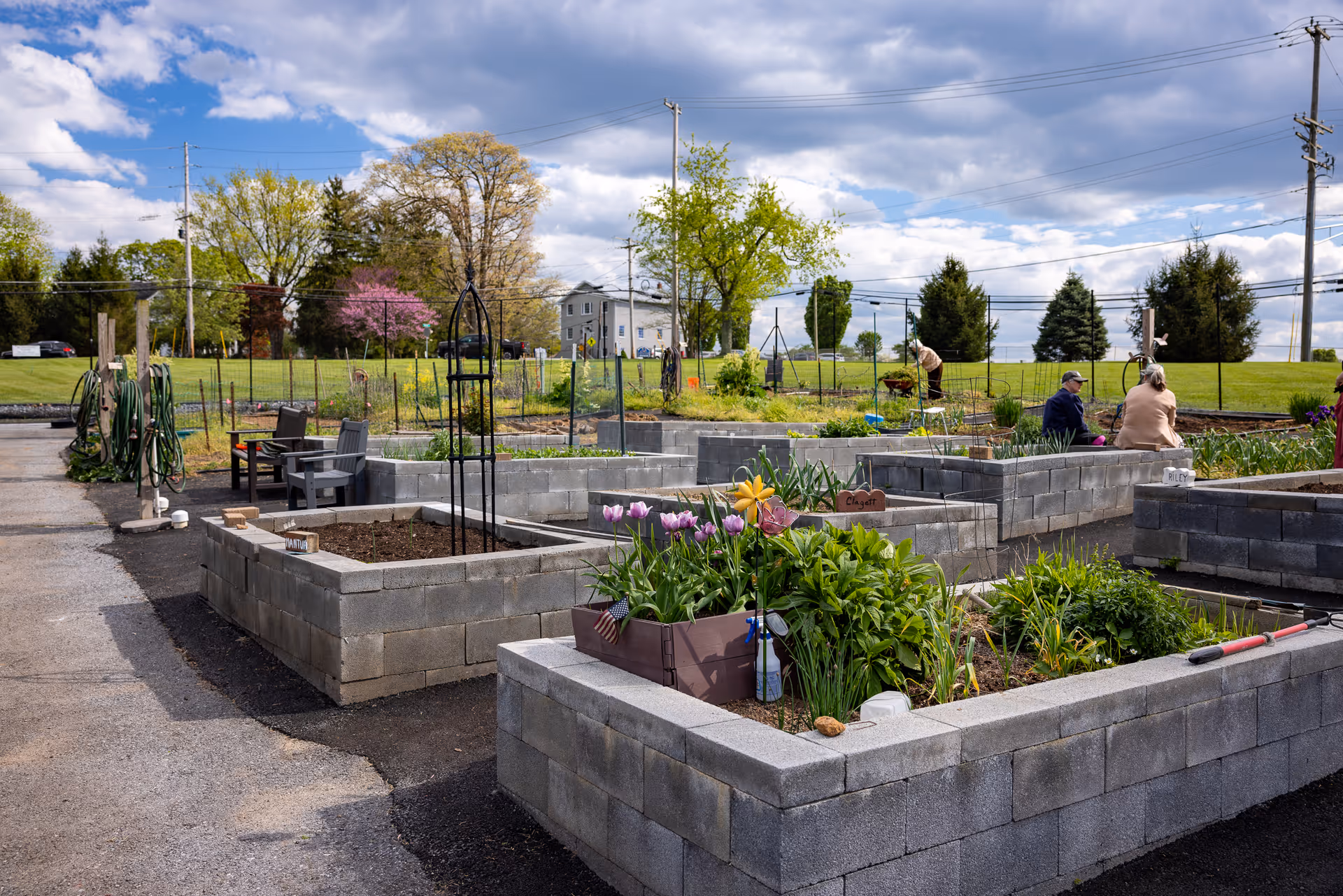 Raised garden beds made of concrete blocks with various plants and flowers growing in them. Several people are tending to the garden in the background under a partly cloudy sky. There are garden hoses and chairs along the paved path beside the garden beds.