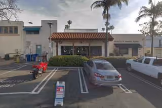 Front exterior view of a single-story building with a tiled roof entrance, palm trees, a red scooter, a gray car, and a white limousine parked in front. There is a signboard on the sidewalk and a cloudy sky overhead.