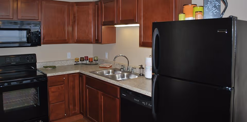 A kitchen area with dark wood cabinets, a black refrigerator, a black stove with oven, a black microwave above the stove, a double stainless steel sink, and a dishwasher. The countertop has a paper towel holder, a soap dispenser, and decorative candles. The kitchen has a neutral wall color and under-cabinet lighting.