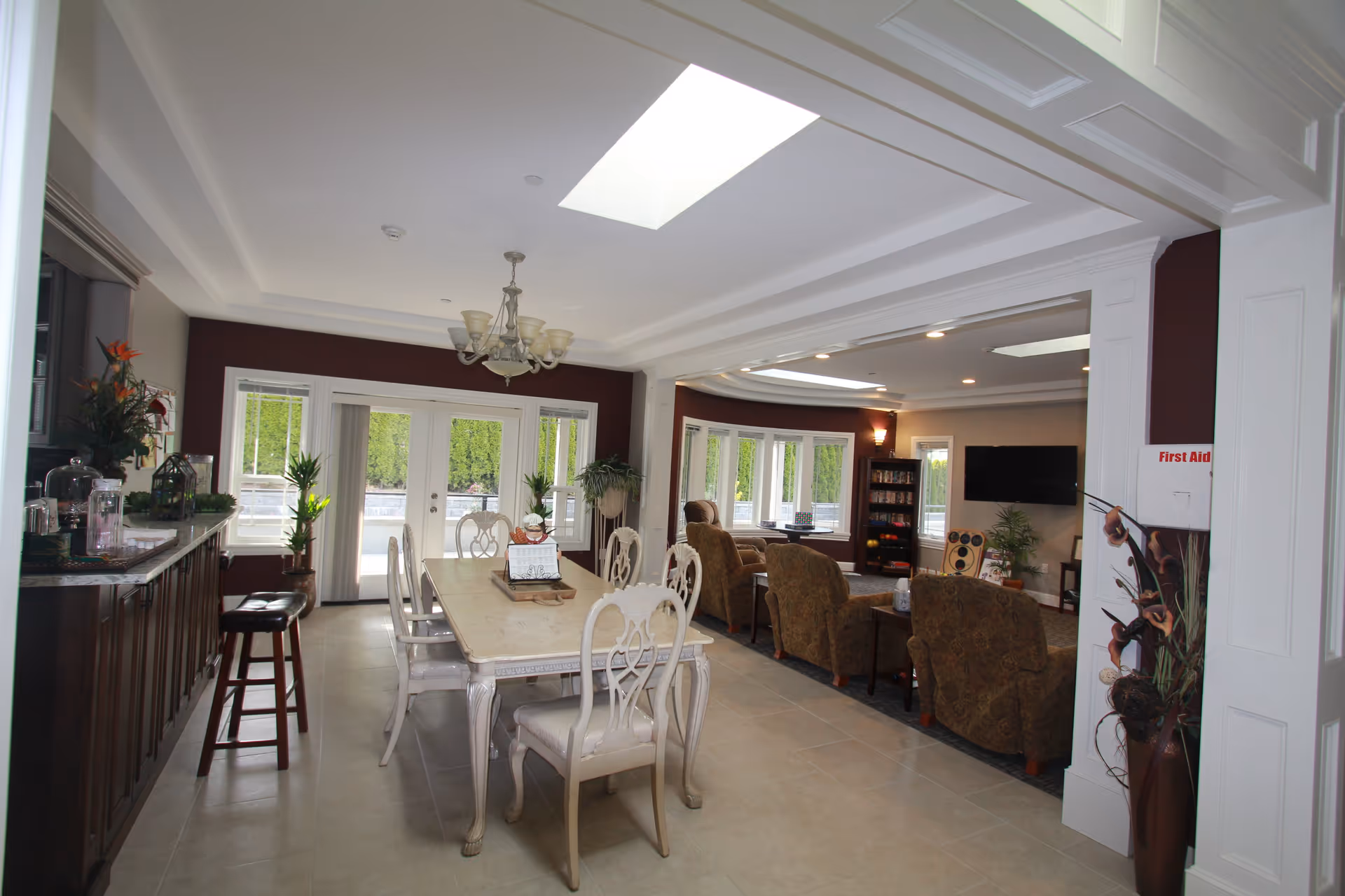 A bright and spacious senior living common area featuring a dining table with six chairs in the foreground and a living room area with four upholstered armchairs facing a wall-mounted TV in the background. The room has large windows and glass doors letting in natural light, a chandelier above the dining table, and a countertop with stools on the left side. There are plants and decorative items placed around the room.