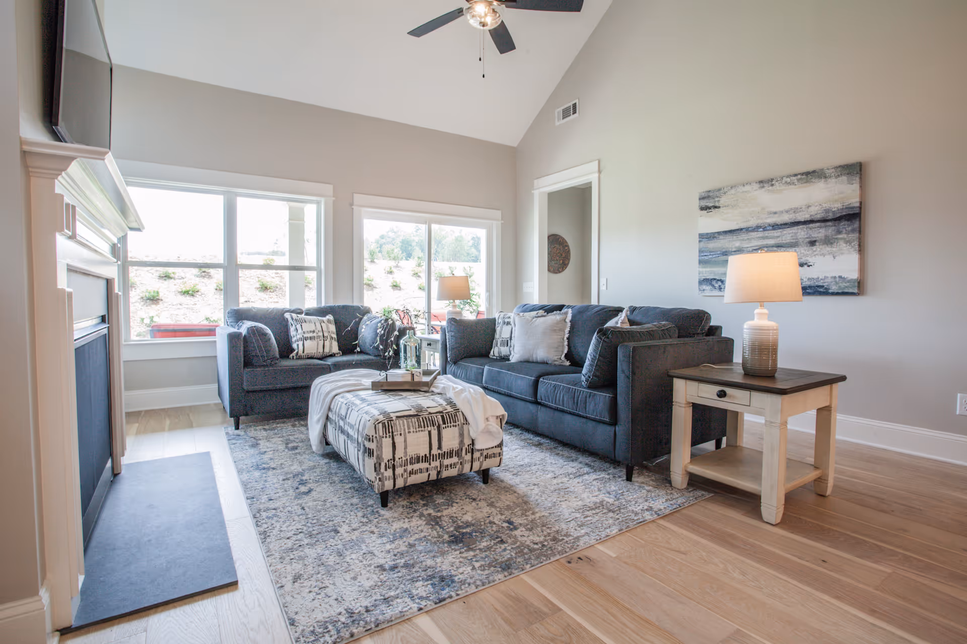 A bright living room with two dark gray sofas, a patterned ottoman, a side table with a lamp, a fireplace, large windows letting in natural light, and a ceiling fan. The room has light wood flooring and neutral-colored walls with a piece of abstract art hanging.