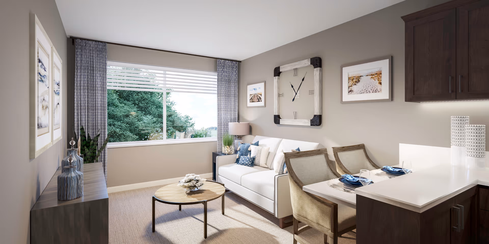 A cozy living room area with a white sofa adorned with blue and white pillows, a round wooden coffee table with a decorative centerpiece, and a side table with a lamp and plant. The room has a large window with blue patterned curtains showing green trees outside. On the wall above the sofa is a large square clock and two framed pictures. To the right, there is a kitchen counter with two place settings and two chairs, and dark wooden cabinets above and below the counter.
