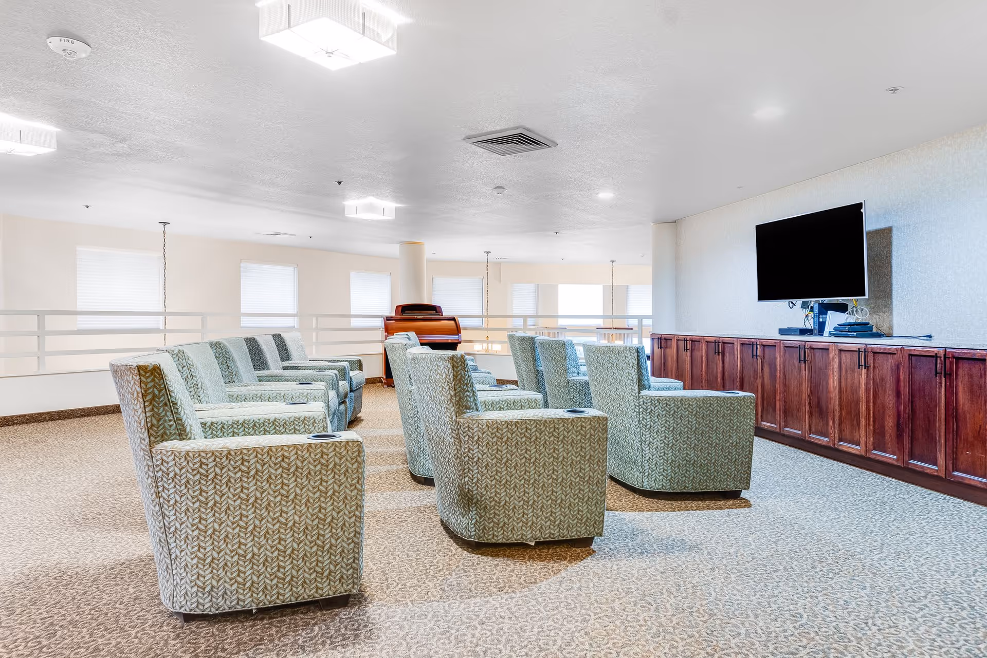 A spacious lounge area with two rows of patterned armchairs facing a wall-mounted flat screen TV above a long wooden cabinet. The room has carpeted flooring, white walls, and several windows letting in natural light.